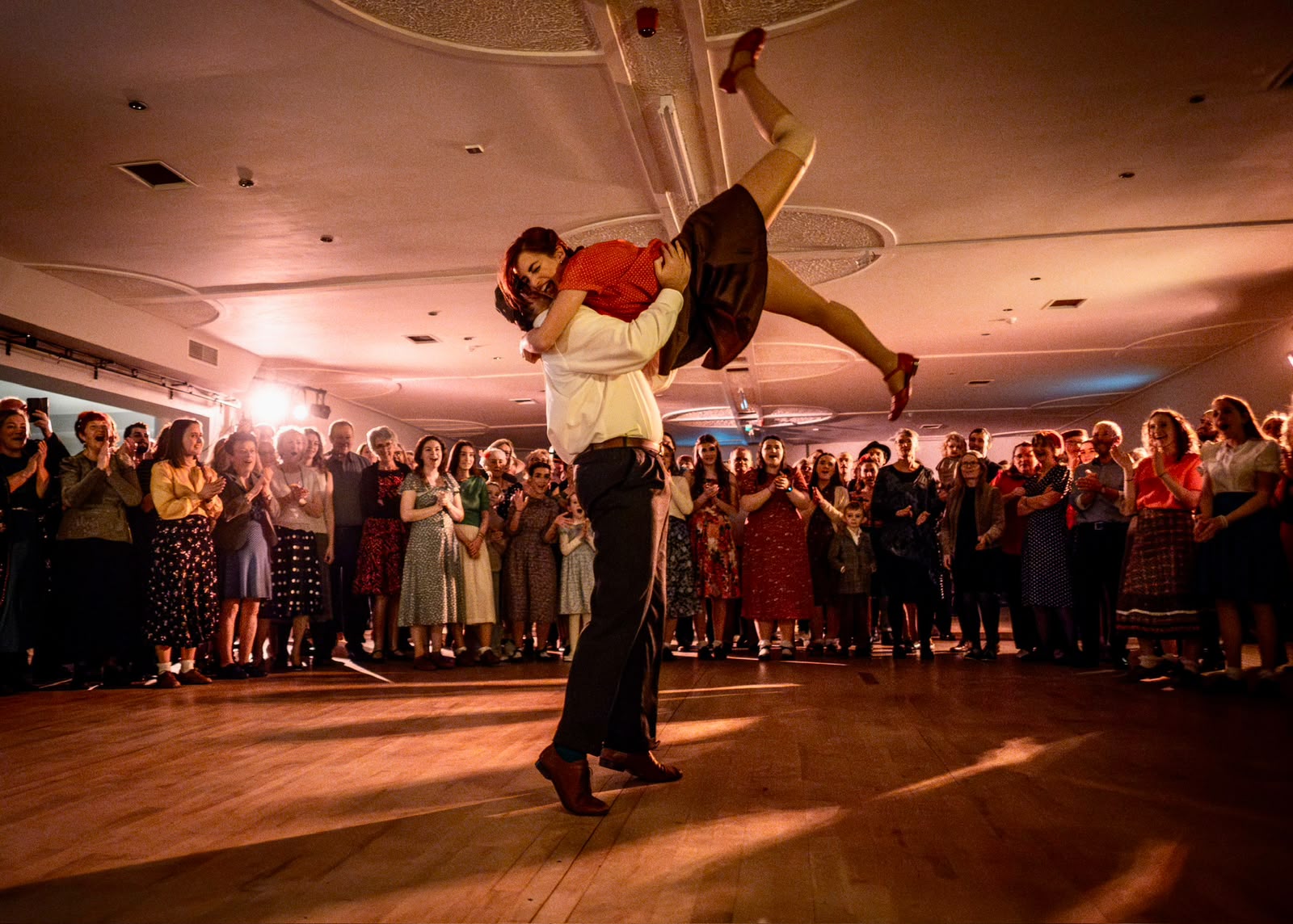 A dancer lifted high overhead on a packed dance hall floor, the crowd ringed around them.
