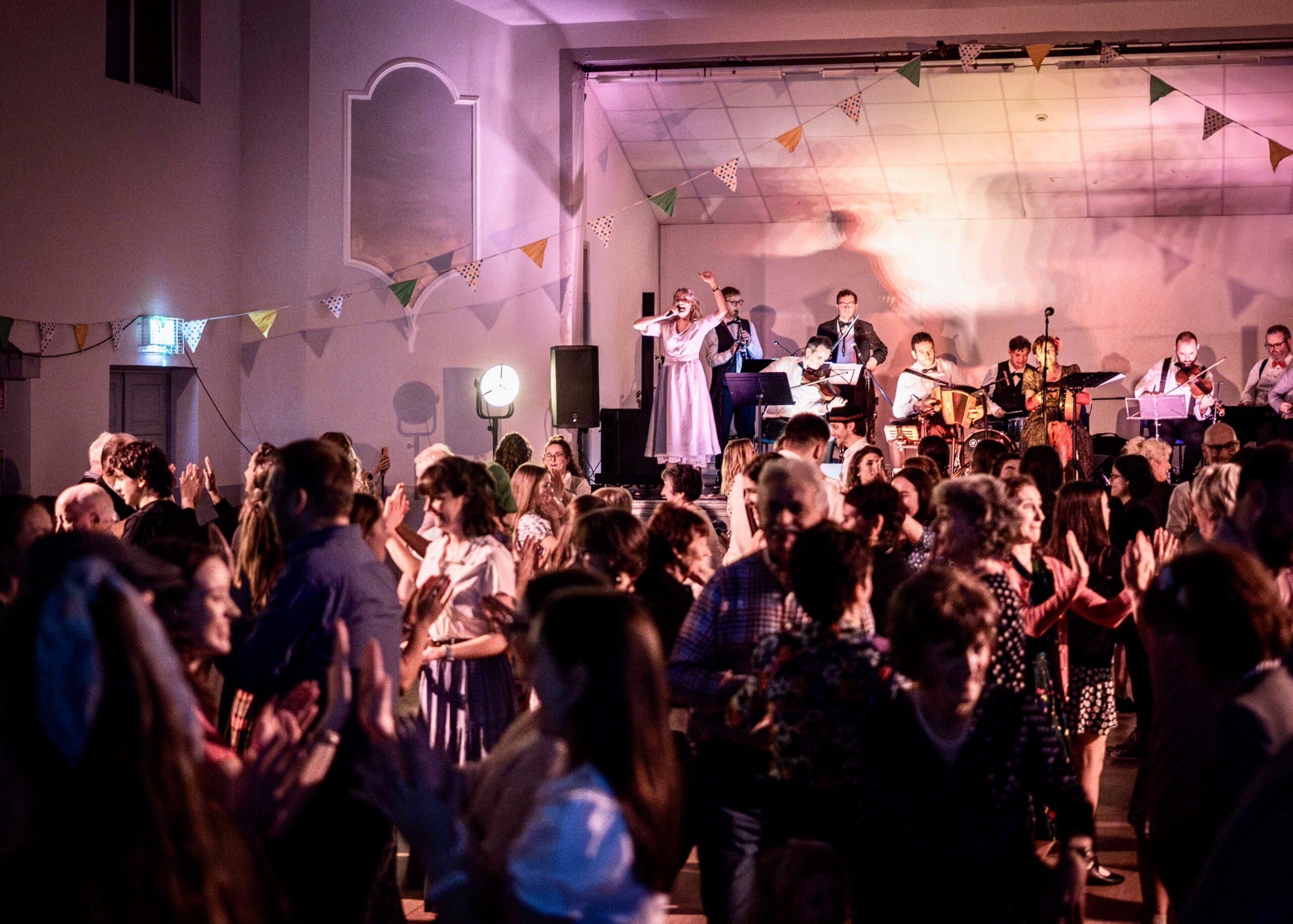 A full hall dancing under coloured lights and bunting, the band playing on the stage behind them.