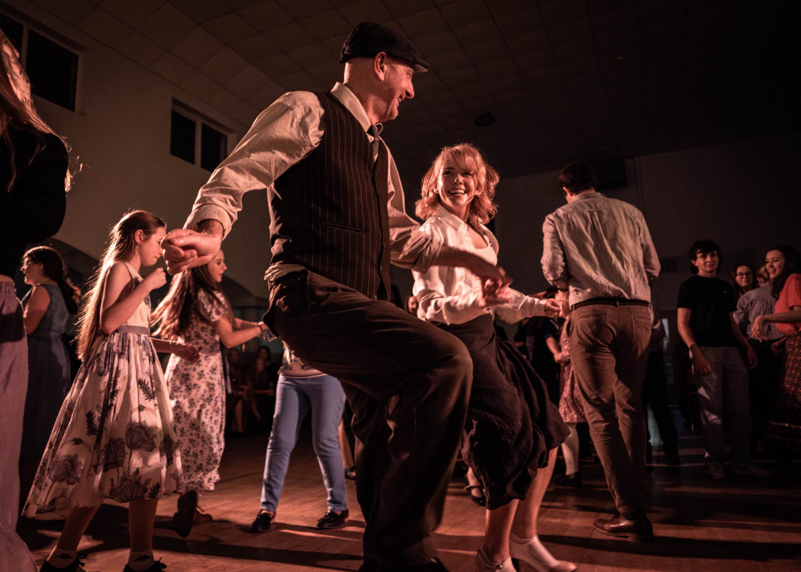 A man in a waistcoat swinging his partner off the ground as children dance beside them.