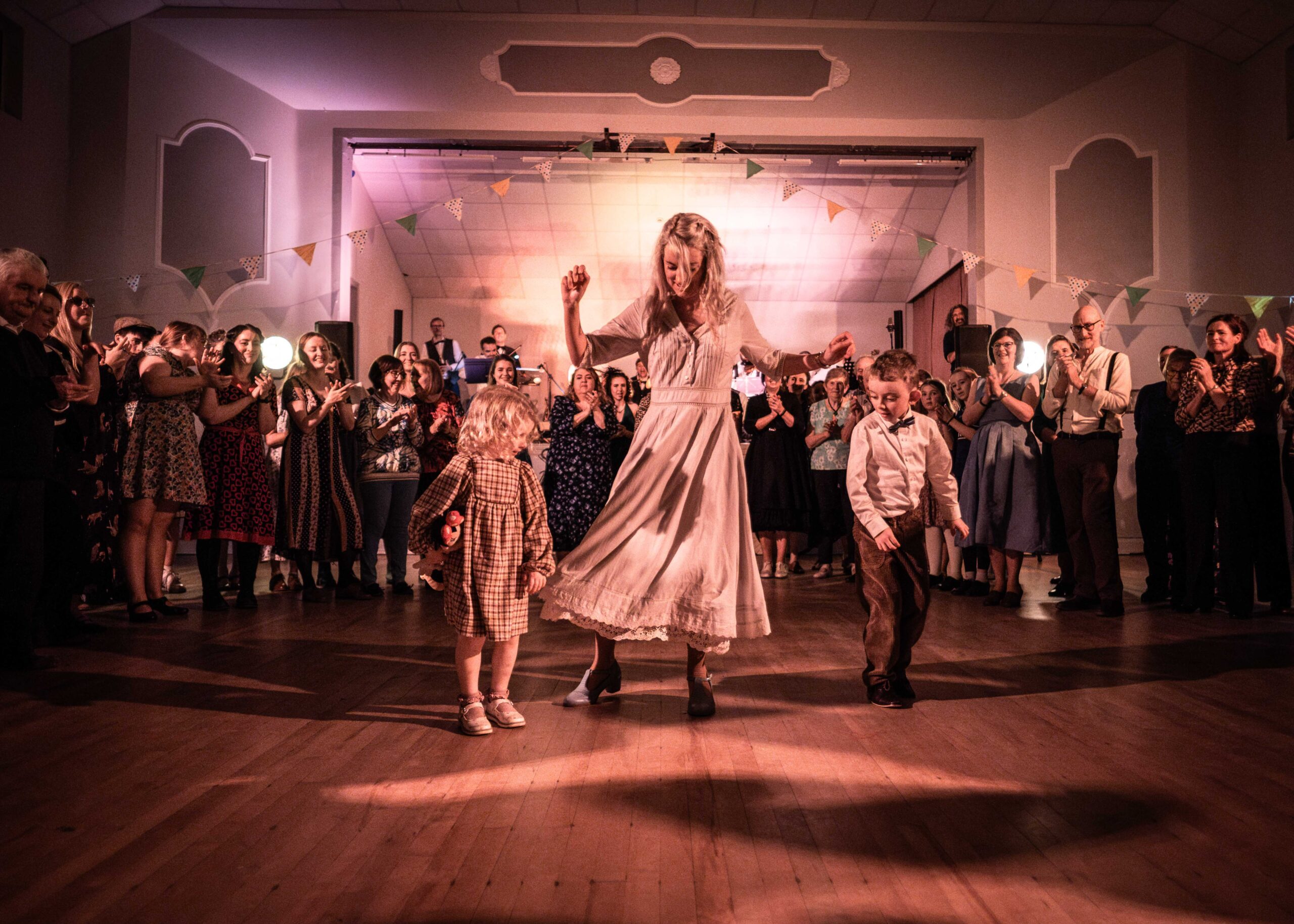 A woman in a cream dress dancing with two small children as the hall claps.